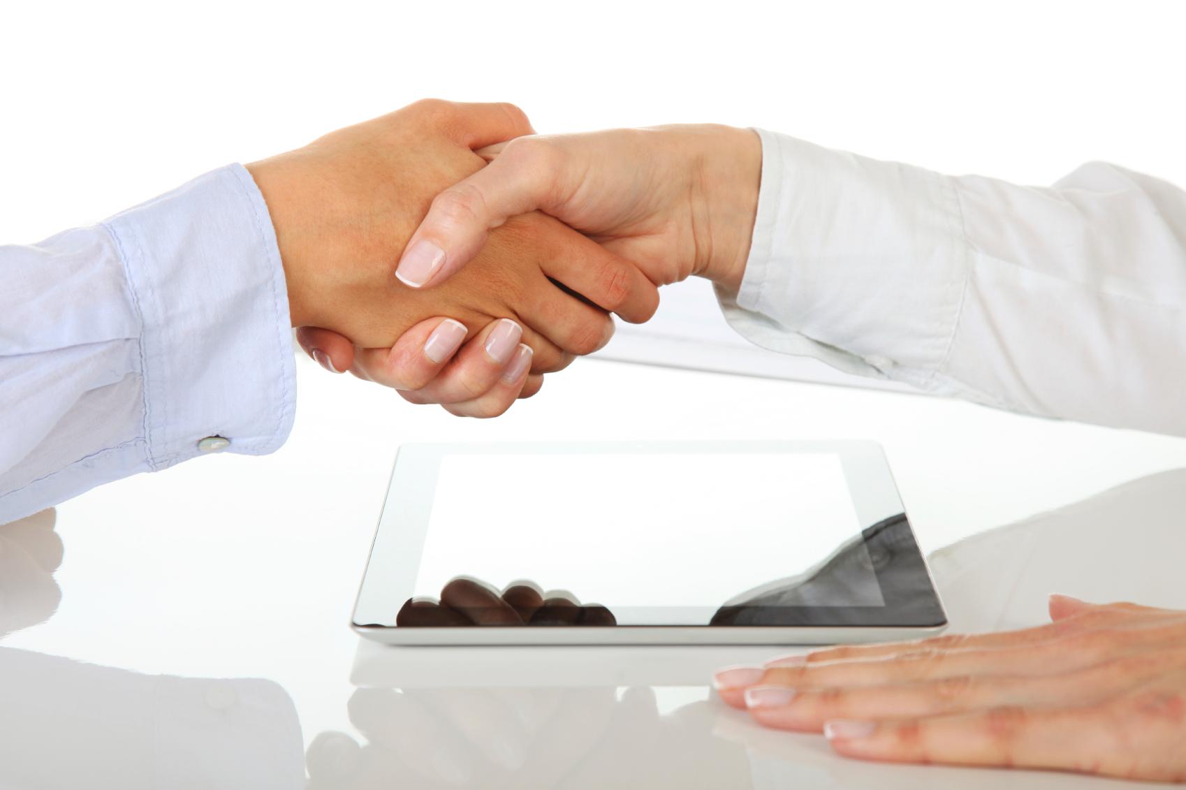 Zwei Personen geben sich über einem Tablet-PC auf einem weißen Tisch die Hand und symbolisieren damit eine Vereinbarung über den Kauf einer Immobilie in Italien