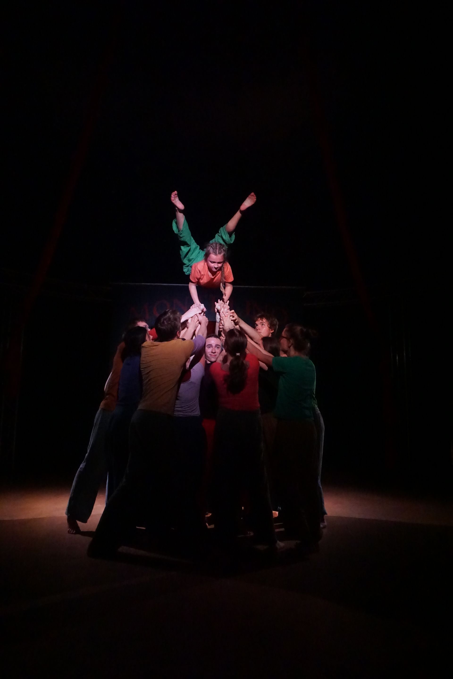 Ein Trainierender wird von vielen dabei unterstützt eine Artistin über sich im Handstand zu halten.