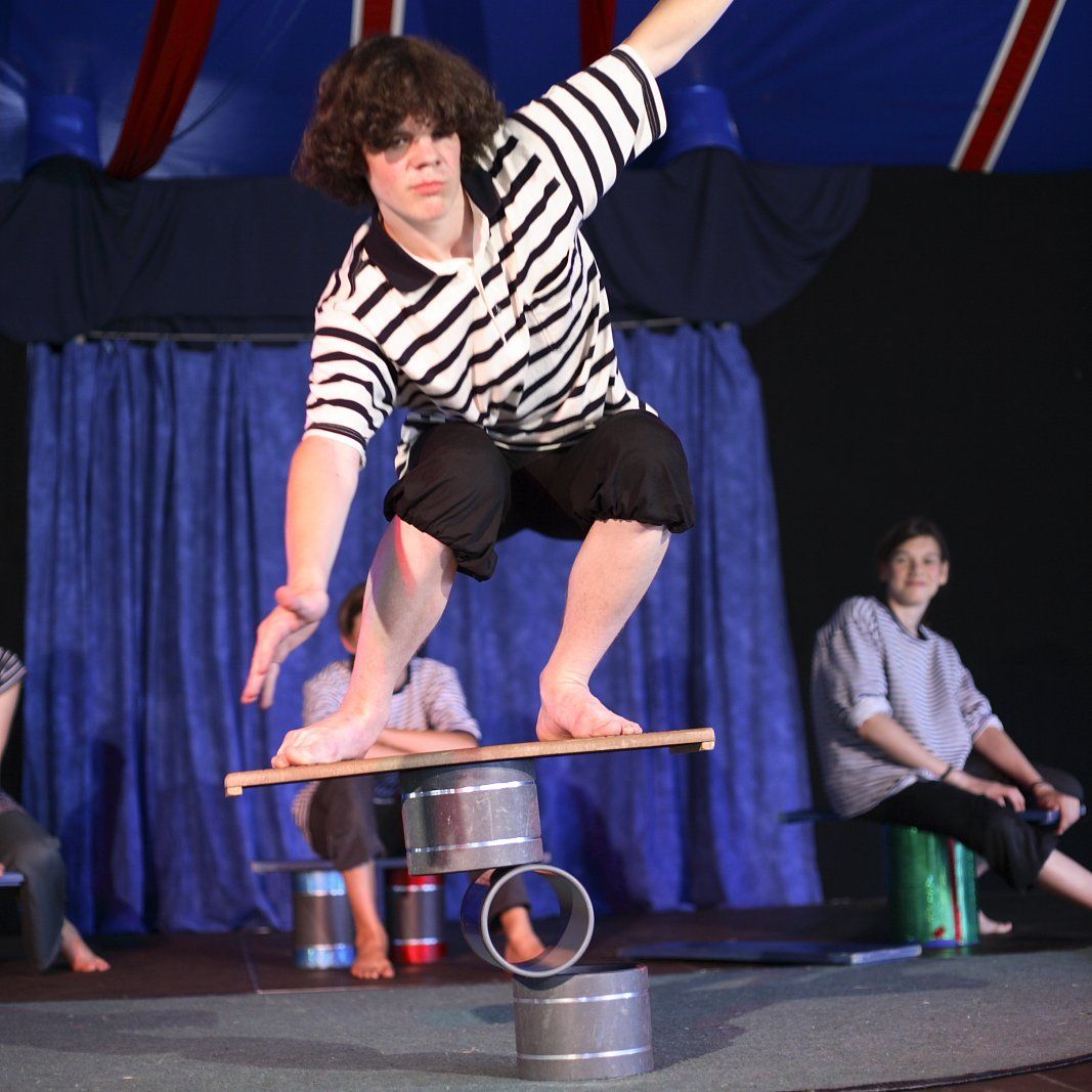 Im Zirkuszelt: Ein Trainierender balanciert auf einem Brett, darunter befinden sich Rollen.