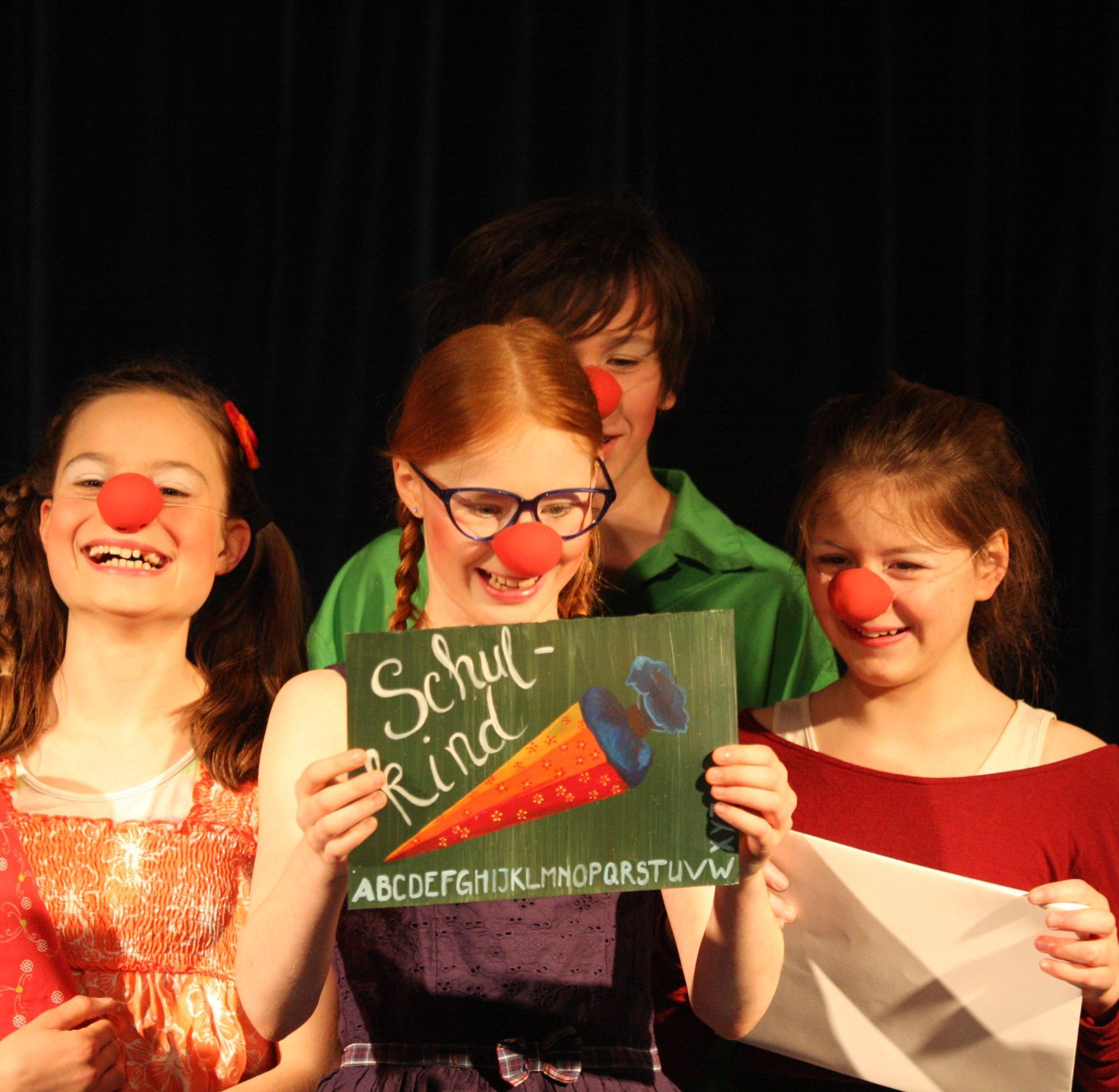 Im Zirkuszelt sind 4 als Clown verkleidete Kinder zu sehen. Eine davon hält ein Schild hoch, darauf ist