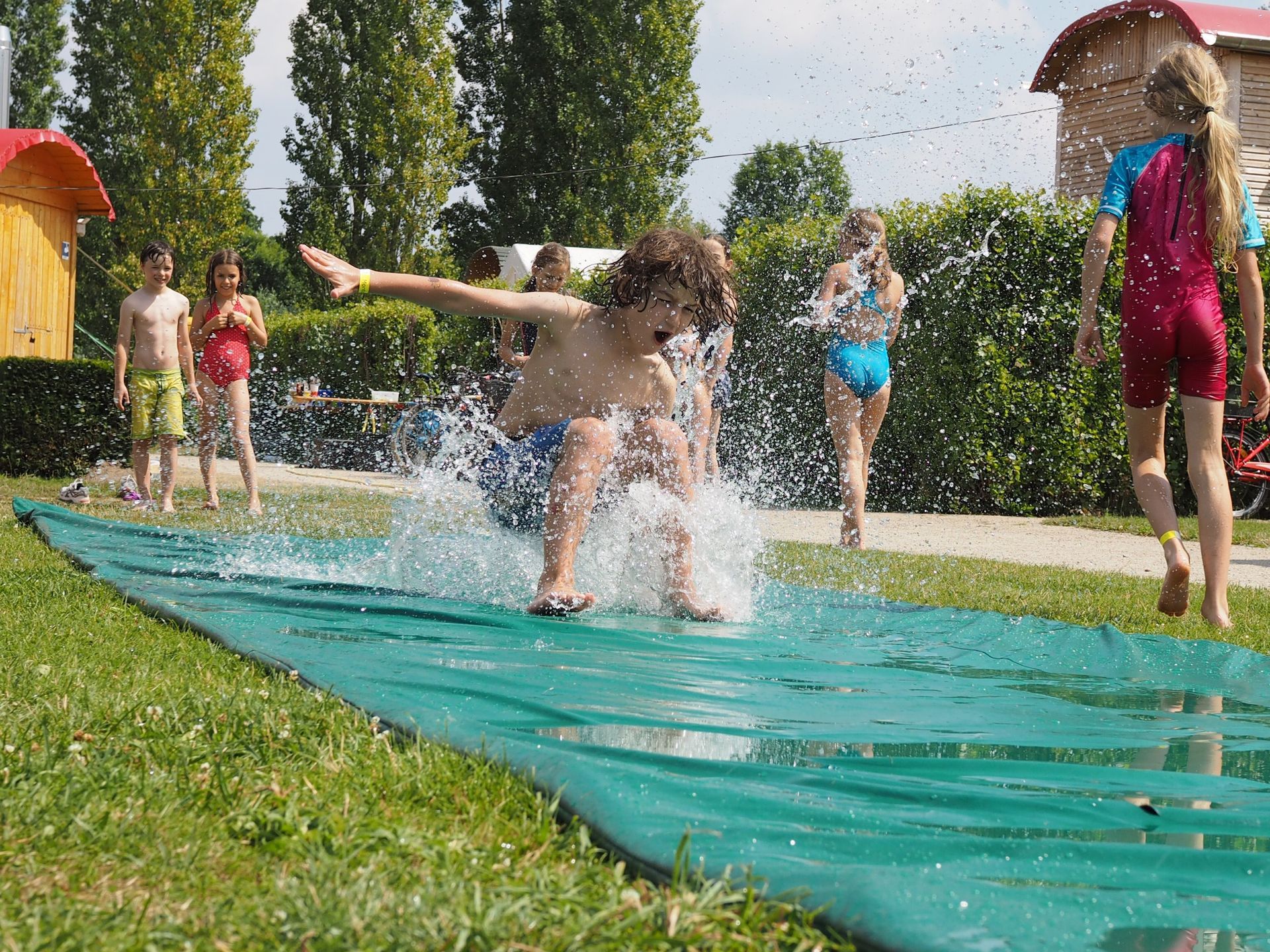 draußen im Park: ein Junge schlittert in Badehose über eine mit Wasser bedeckte Plane. Er hat Spaß. Ferienbetreuung.