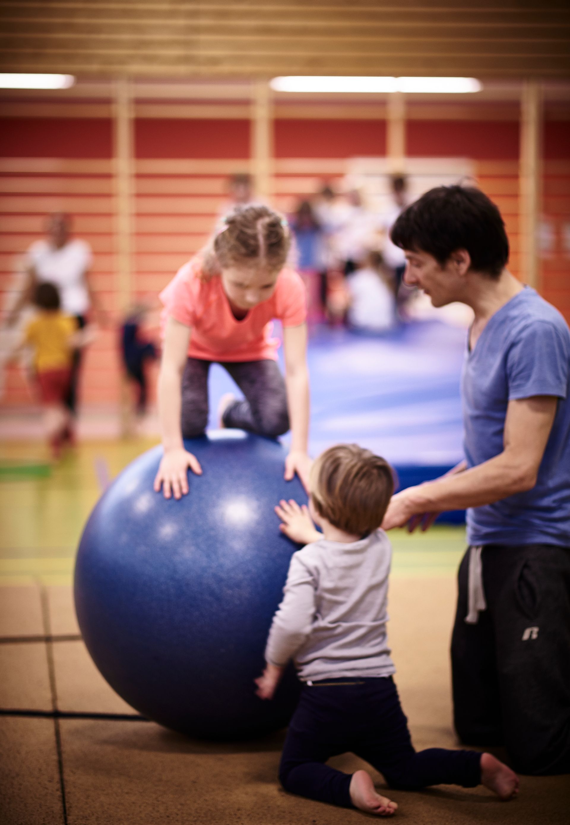 Zu sehen sind 2 Kinder und ein Trainer. Ein Kind sucht Halt auf einer großen Kugel. Der Trainer und das 2. Kind halten die Kugel fest.