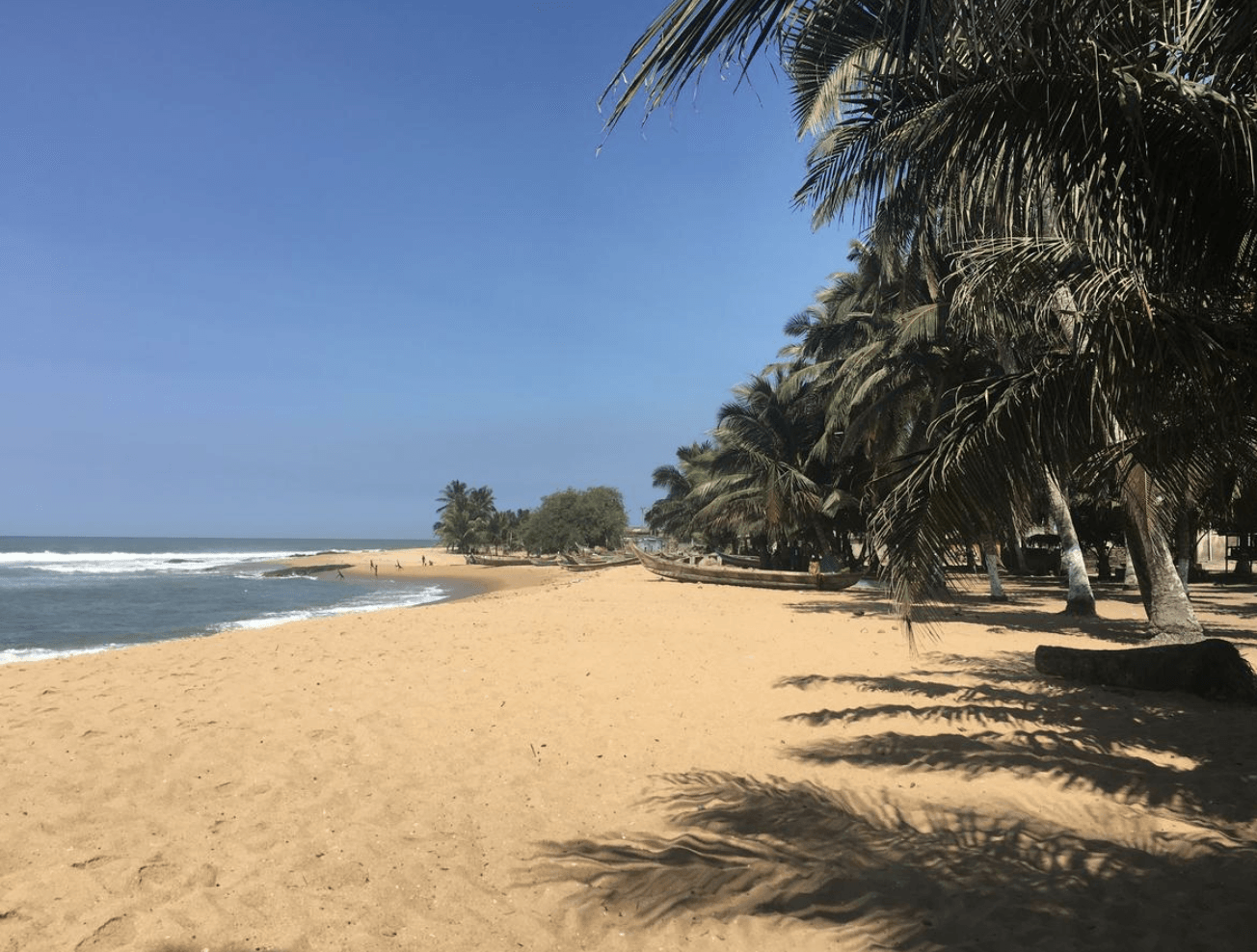 Palmen an einem goldgelben Strand, Meer mit kleineren Wellen. Im Hintergrund Boote der Einheimischen und Menschen