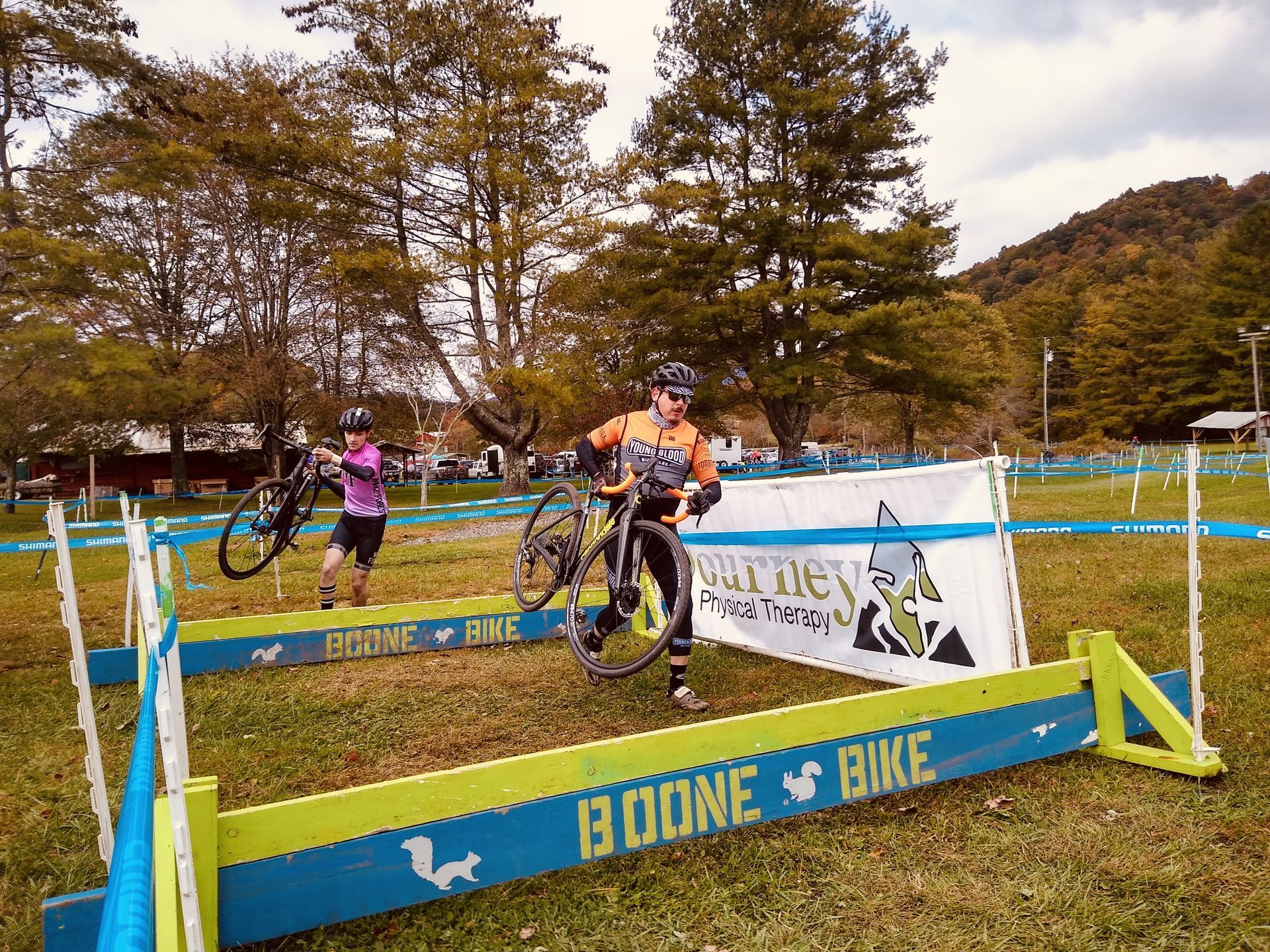 two young men are hold drop bar bikes while running over small barriers in a grassy area. There are plastic stakes with event tape to mark a course.