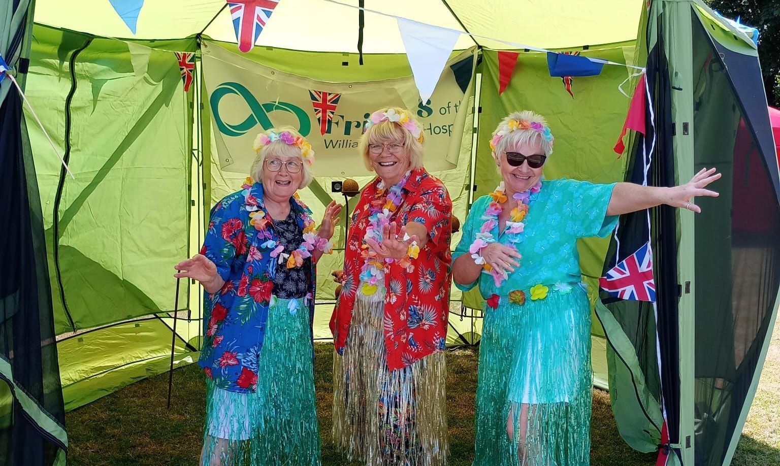 Three smiling women in Hawaiian shirts standing in a green tent with bunting and a sign saying Friends of William Harvey Hospital behind them
