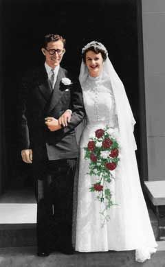 Smiling bride and groom standing arm in arm, bride holding red and white flower bouquet.