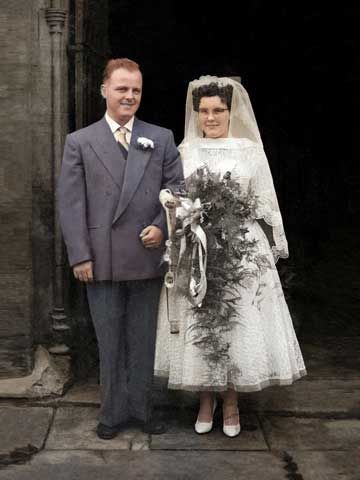 Bride and groom posing outside a doorway, bride in lace gown holding a bouquet, colour photo.