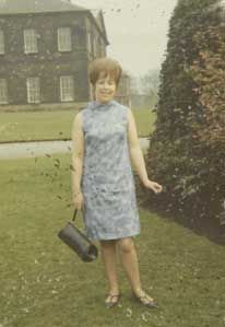 Woman in a sleeveless blue dress smiles while posing on grass in front of a large stone building and garden. Basic Scan.