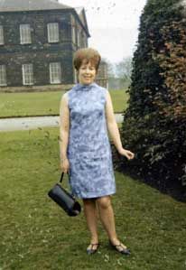 Woman in a sleeveless blue dress smiles while posing on grass in front of a large stone building and garden. Standard Scan.