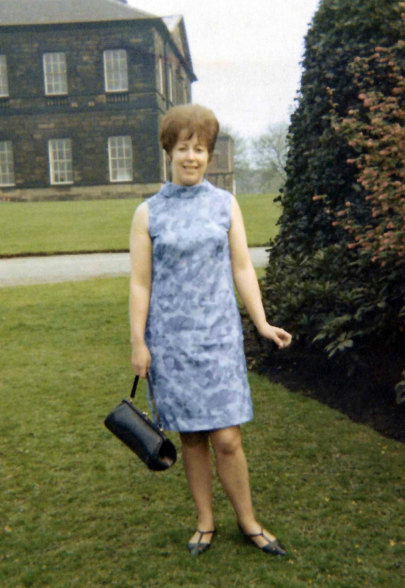 Woman in a sleeveless blue dress smiles while posing on grass in front of a large stone building and garden. Premium Scan.