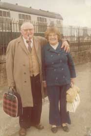 Elderly couple smiling at a train station, the man with a pipe and plaid bag, the woman holds a white handbag. Basic Scan.