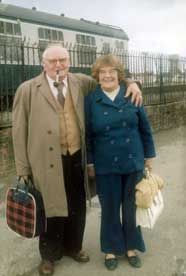 Elderly couple smiling at a train station, the man with a pipe and plaid bag, the woman holds a white handbag. Standard Scan.