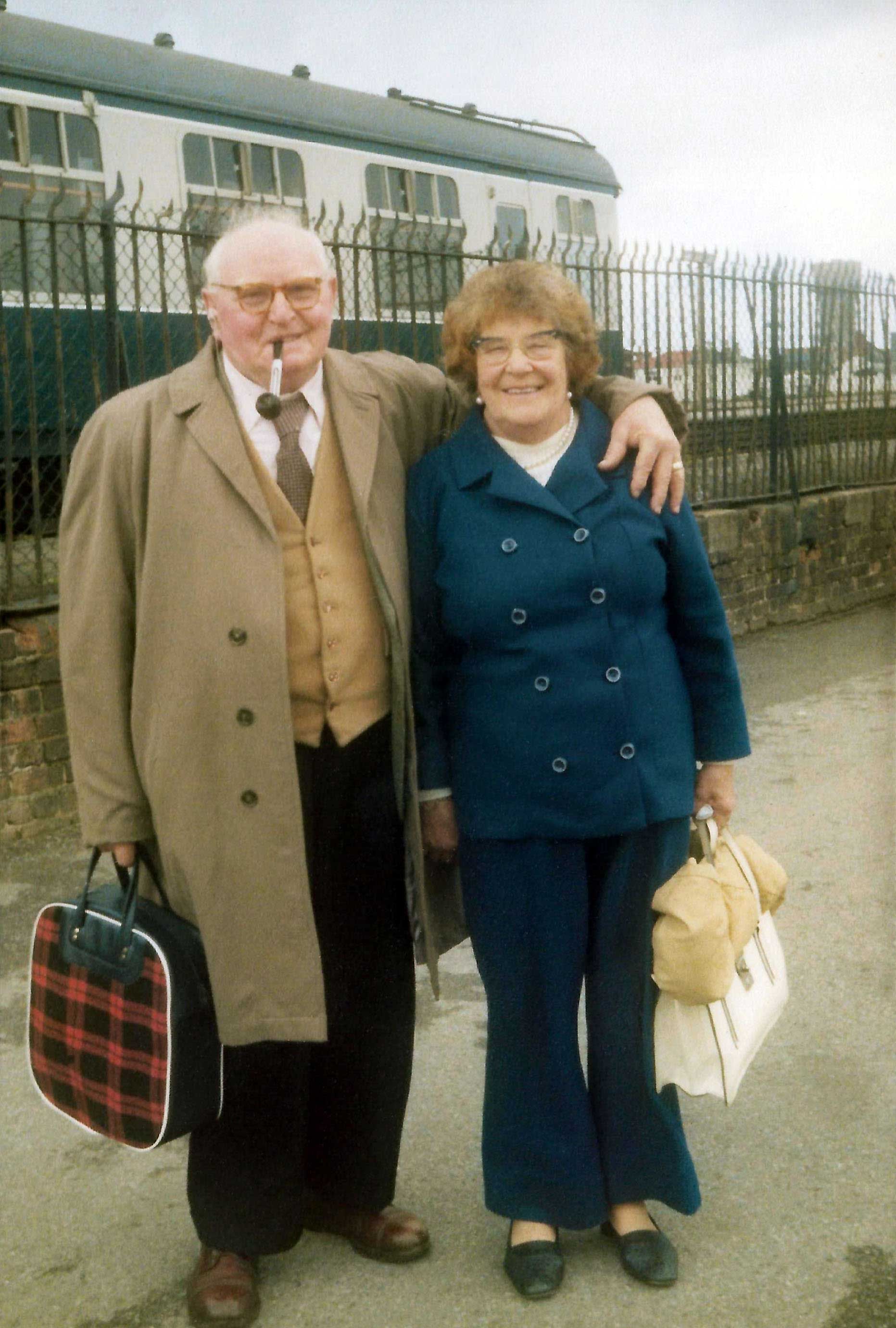 Elderly couple smiling at a train station, the man with a pipe and plaid bag, the woman holds a white handbag. Premium Scan.