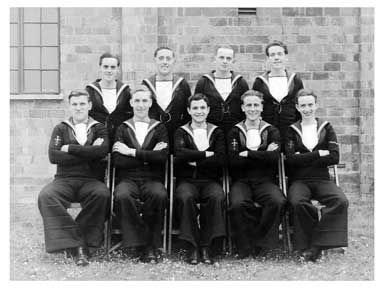 Black and white photo of nine sailors in uniform posing against a brick wall, five seated and four standing