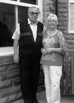 Black and white photo of an elderly couple standing together outside a stone building.