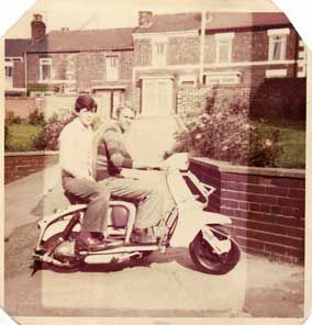 Faded photo of two lads on a scooter in front of houses.