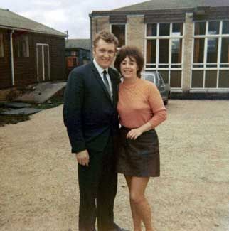 A smiling couple poses outside in 1960s attire, standing near buildings and a vintage car in the background.
