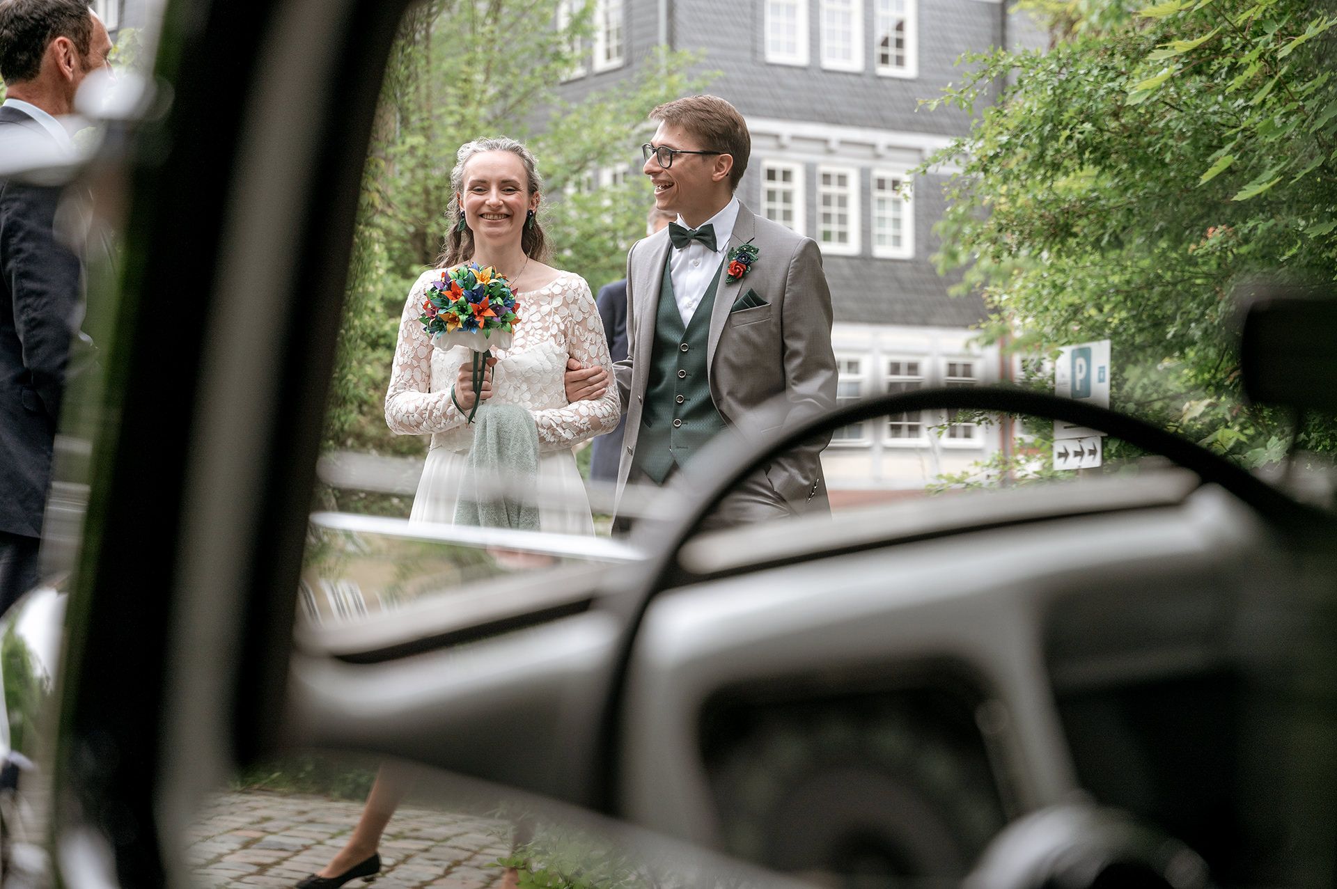 Durch die Frontscheibe eines Oldtimers sieht man ein glückliches Brautpaar, das in ihrem Hochzeitsoutfit posiert.