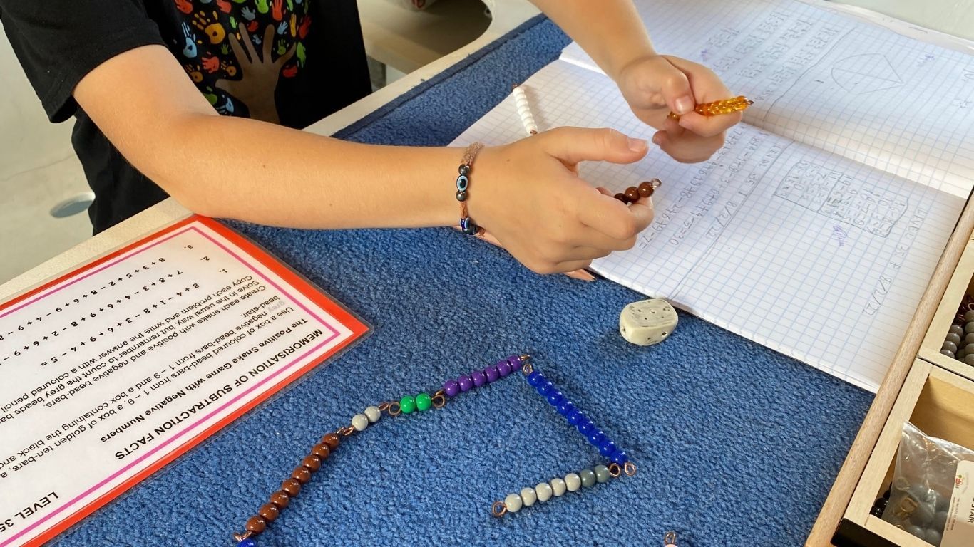 A child's hands can be seen holding and counting coloured bead-bars above a mat with an exercise book and more beads.