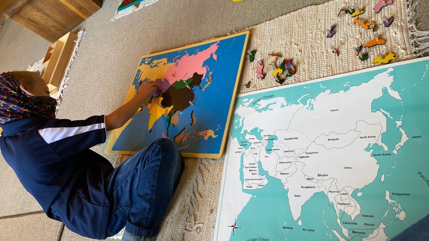A child placing the puzzle piece of a country into the puzzle map of Asia. There are pieces next to the map, scattered on the mat.