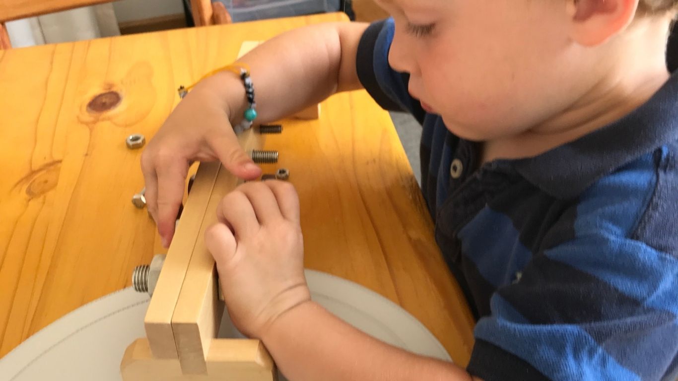 A young child attempts to undo a set of nuts and bolts in a wooden beam that rests on a table.