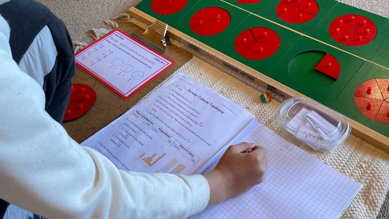 Ein Schulheft liegt aufgeschlagen auf einer Matte. Daneben liegen die Übungskarte und ein Satz roter und grüner Brucheinsätze. Man sieht die Hand eines Kindes, das die Arbeit aufzeichnet.