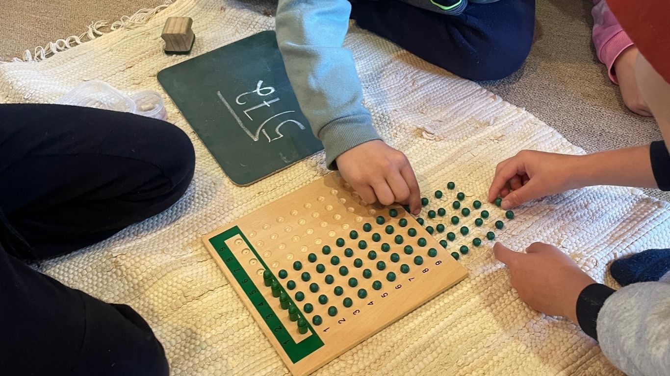 A long division problem is being solved with the Montessori division board (green unit beads are being shared out).