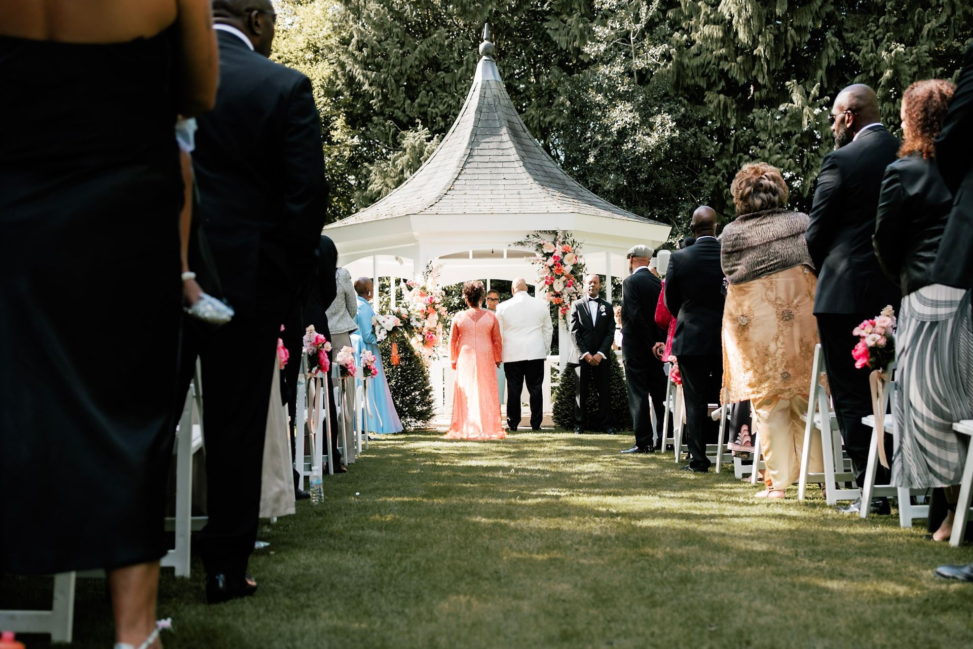 Bride in orange wedding dress and groom dressed in white tuxedo holding hands and smiling with bac outside the Orangery Maidstone gazebo with guests seated outside