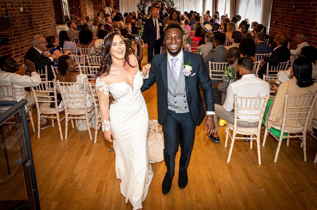 White bride and black groom smiling with their wedding guests behind them as they walk up the aisle