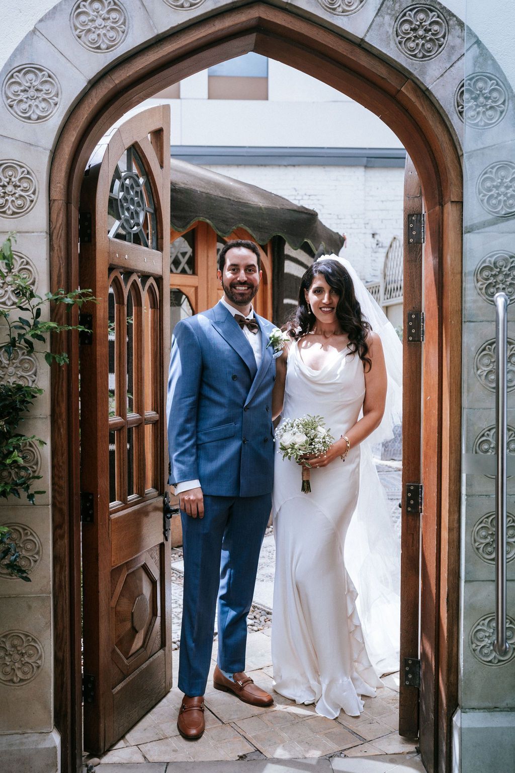 Bride in white dress holding flower bouquet standing with groom in blue dress by a door arch