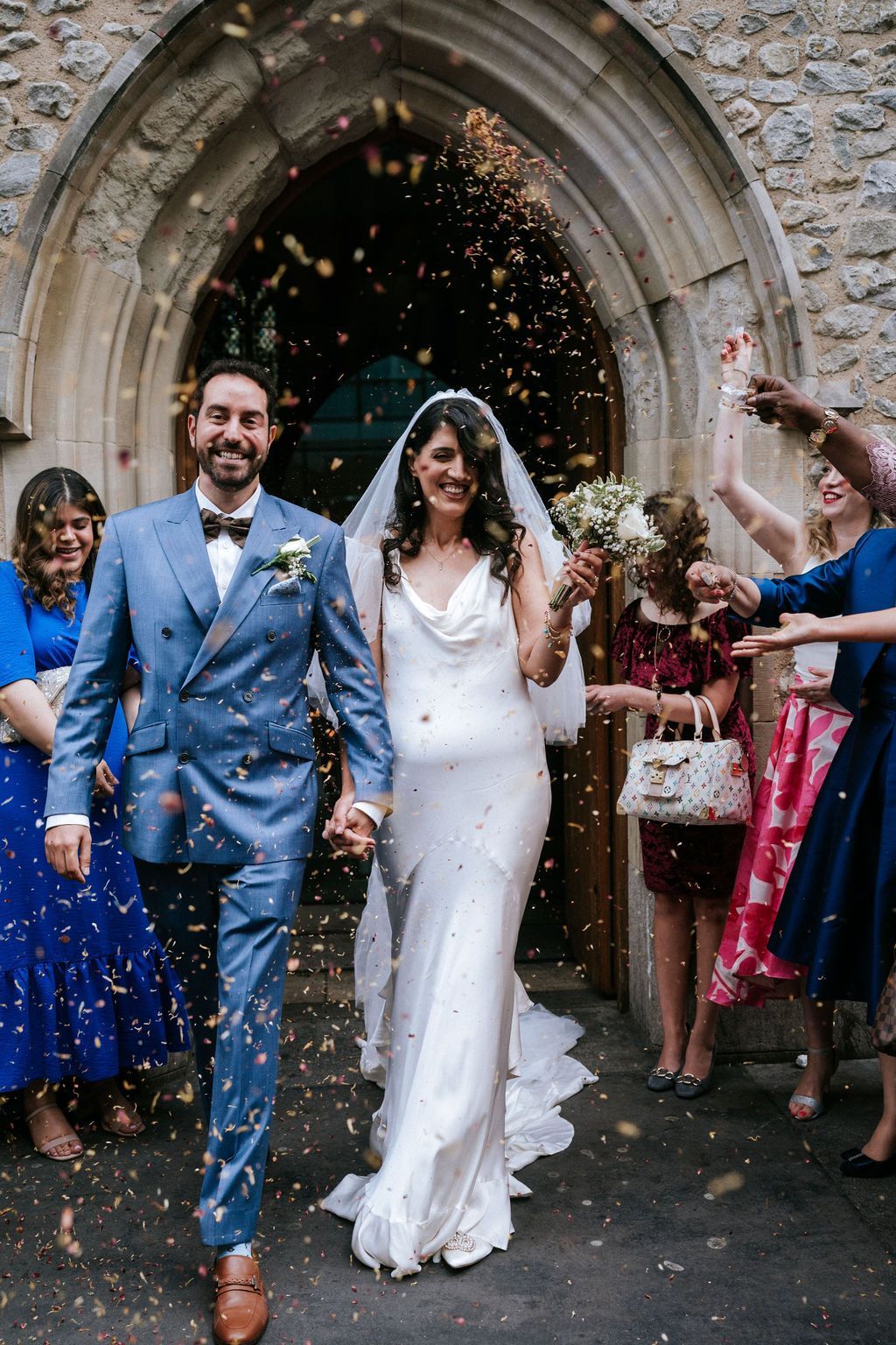Bride and groom holding hands leaving church and guests throwing confetti