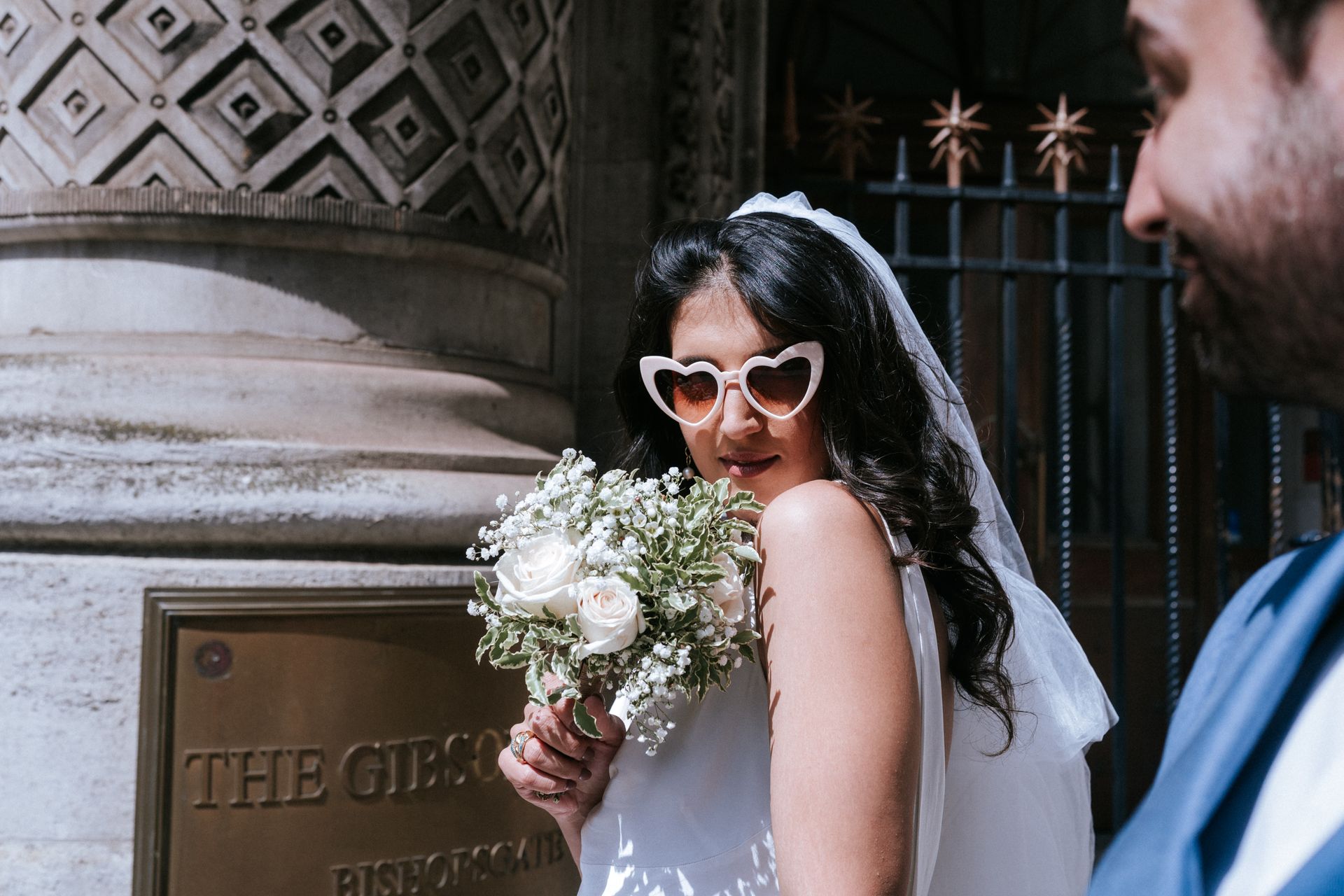 WHite woman in wedding dress with heart shaped sunglasses on and holding up bouquet