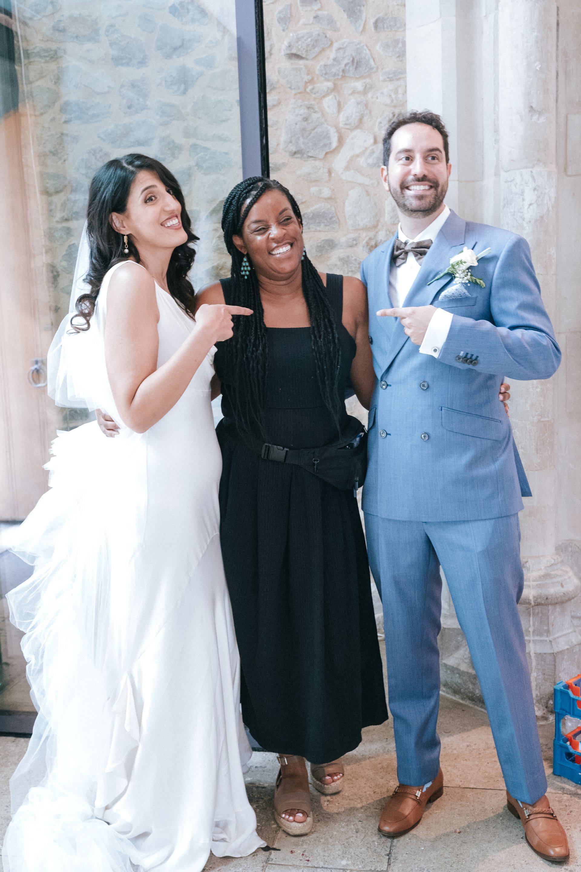 Bride in white dress and groom in blue suit standing with black woman wedding planner and pointing