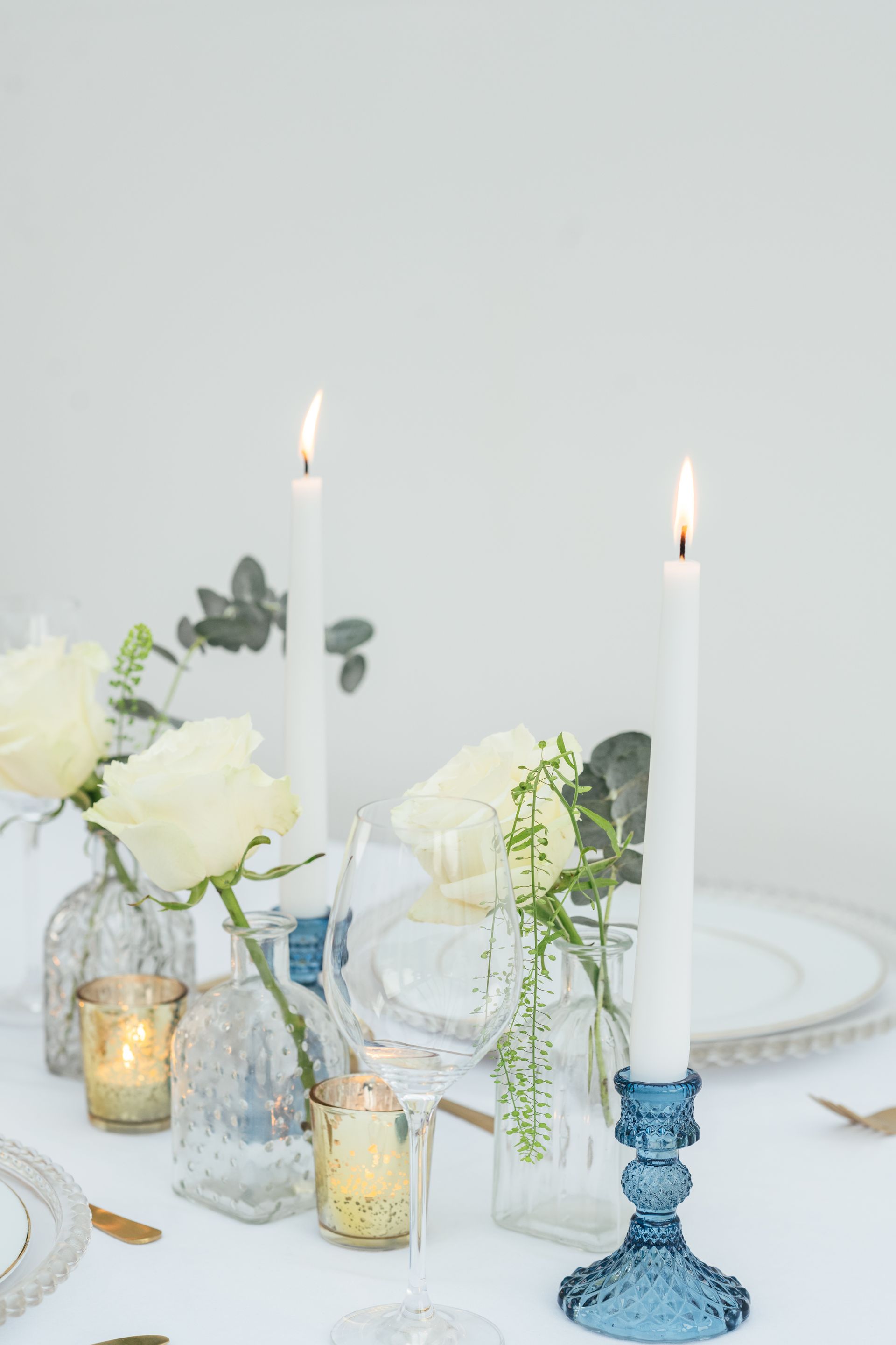 Blue glass candlestick with white candles on a table with cream roses in bud vases