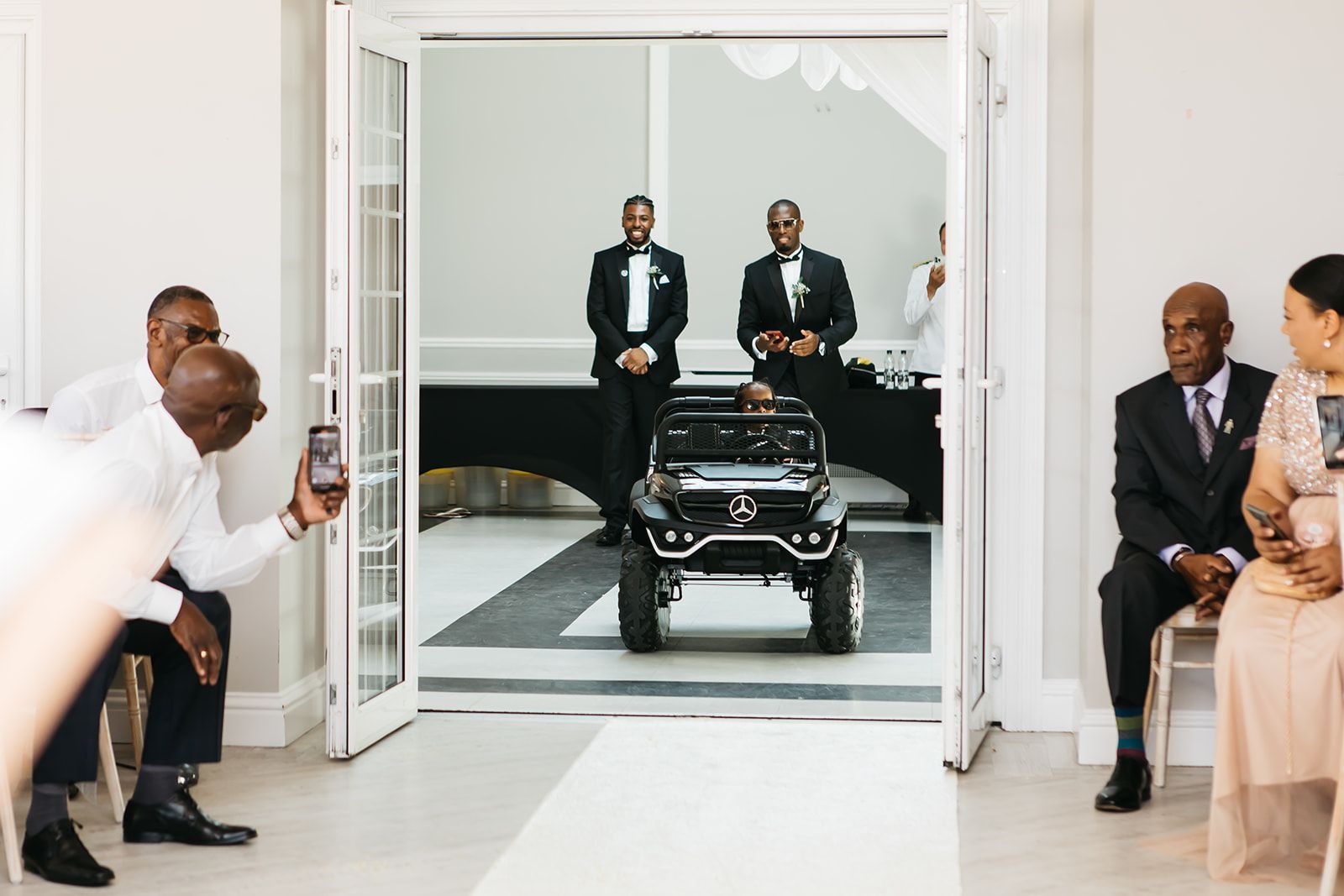 A young black boy in a black suit sitting behind a mini black Mercedes and driving it down the aisle