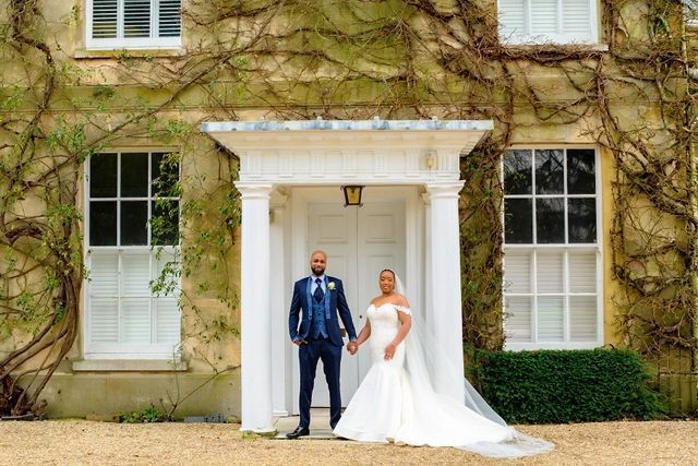 Black bride in white long dress and black groom in blue suit holding hands and standing outside Northbrook Park house entrance