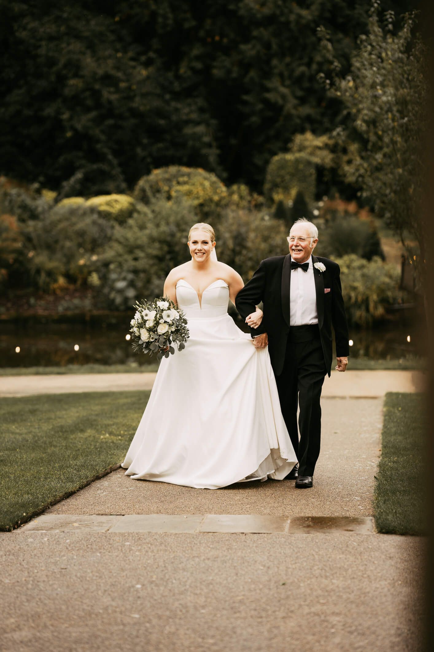 White bride walking on a path with her father who is in black tuxedo