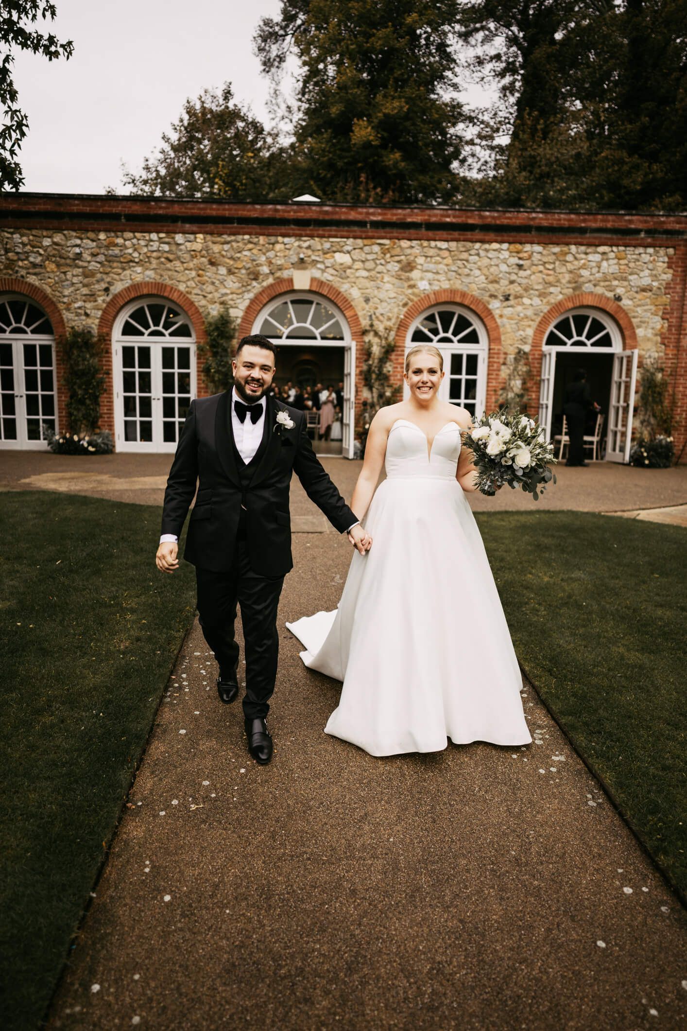 Bride in white sleeveless wedding dress and groom dressed in black tuxedo holding hands and smiling outside the Orangery Maidstone