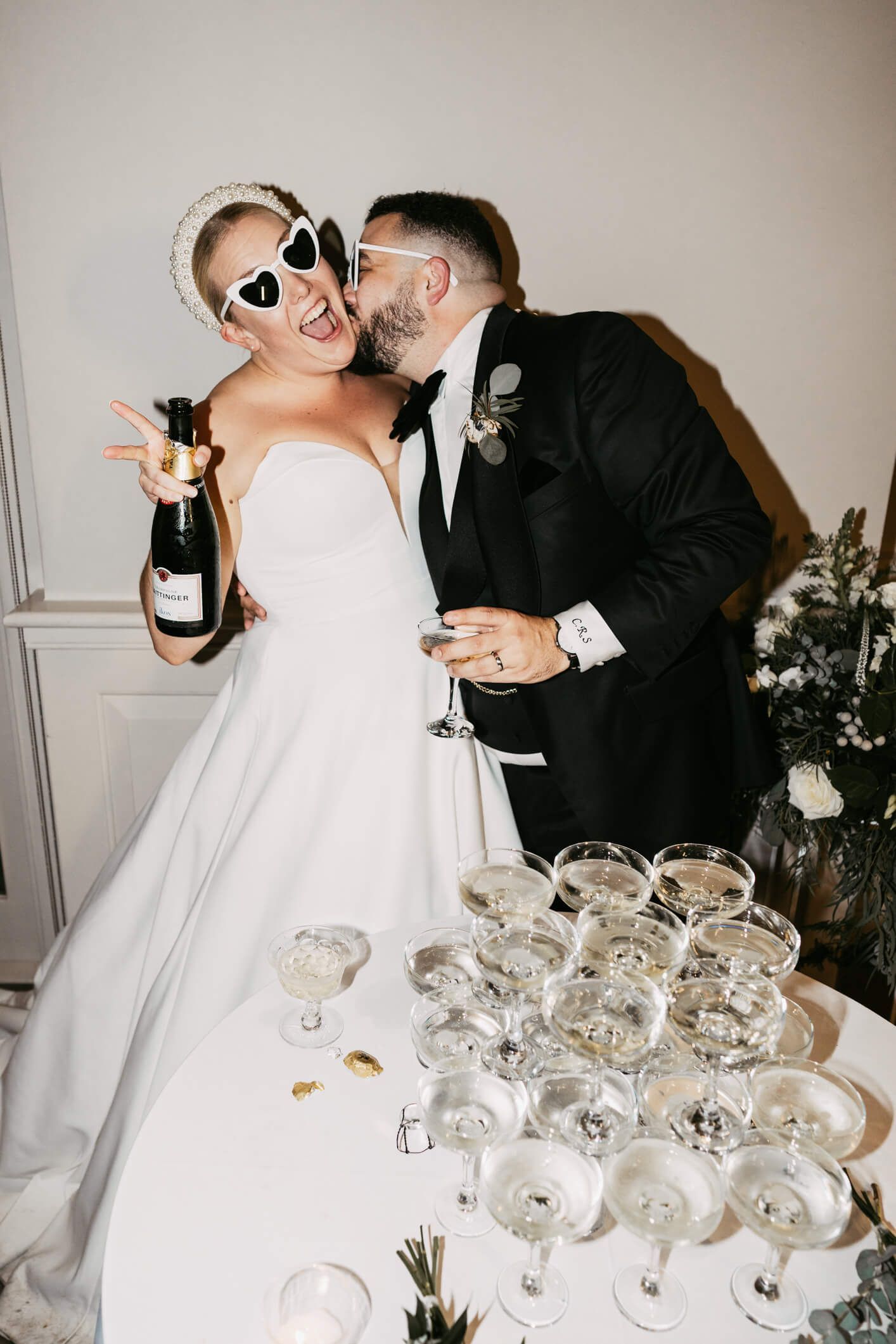A white bride and groom standing near their champagne tower. Groom kisses bride's neck and the bride laughs whilst holding a bottle of champagne