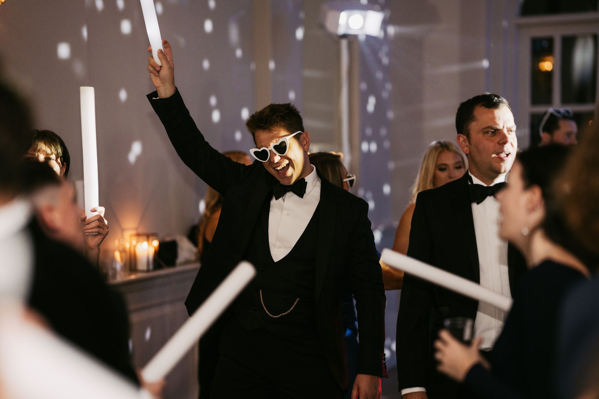 White man in black tuxedo dancing and holding up a white glow stick and wearing white heart sunglasses