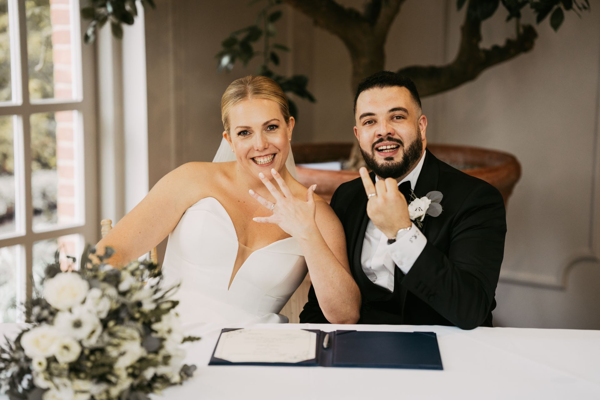 White bride and groom smiling to the camera and holding up their fingers with wedding rings on.
