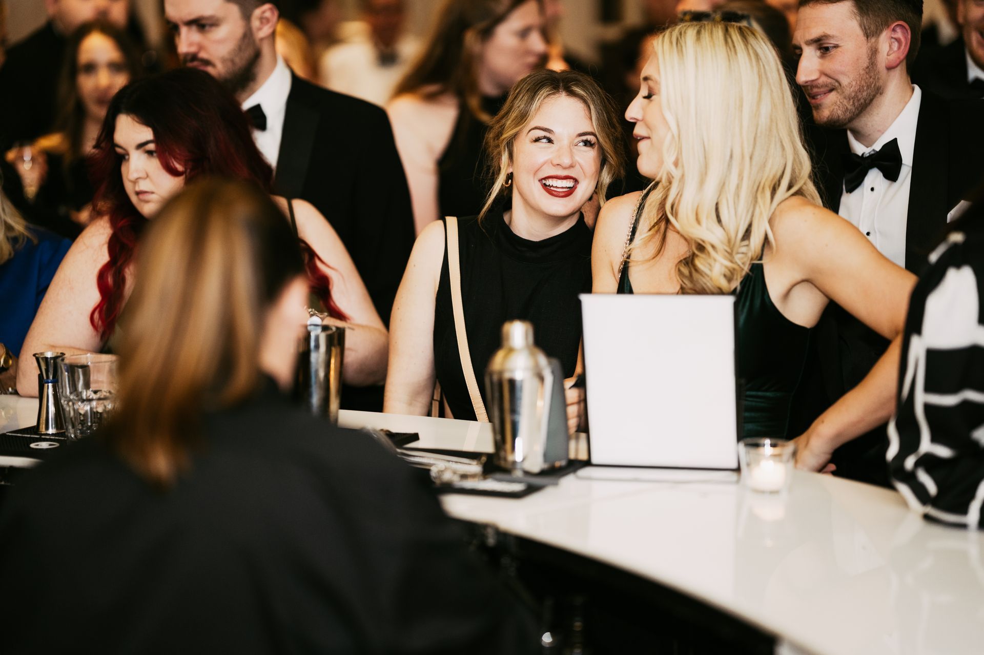 Blond women in black dresses smiling at each other and leaning on a white bar