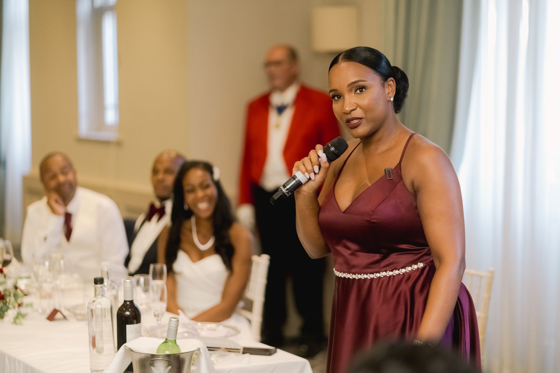 Black bridesmaid in deep red dress standing and giving a speech as bride and groom laugh