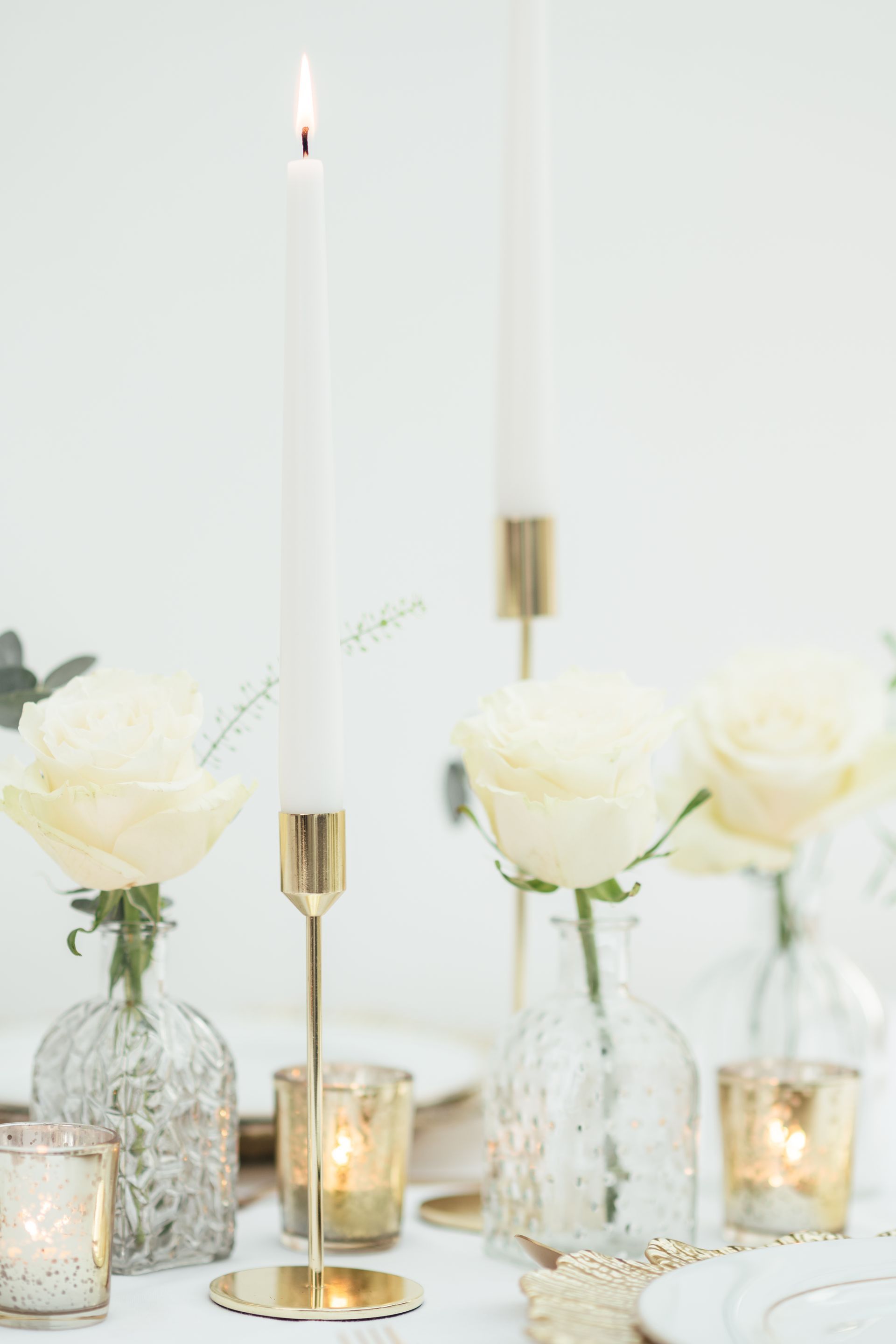 Gold candleholders with white candle displayed on a table with gold tea lights and cream roses in bud vases
