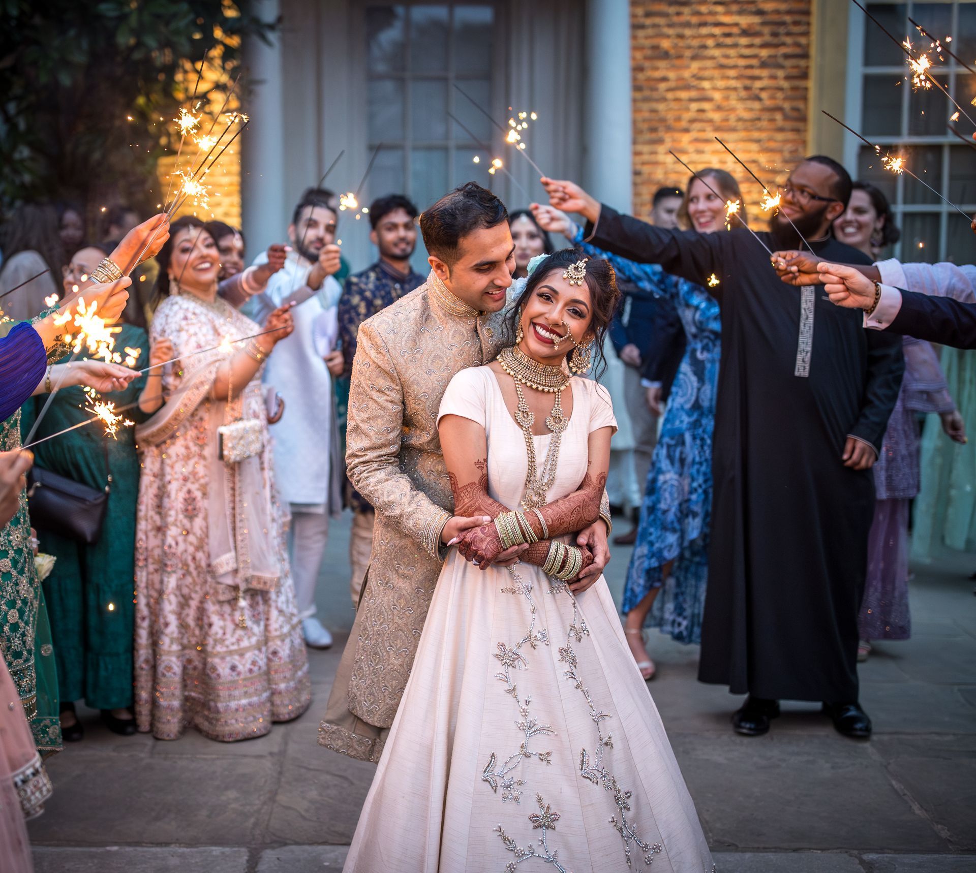 Indian bride and groom facing the camera and hugging as guests raise lit sparklers