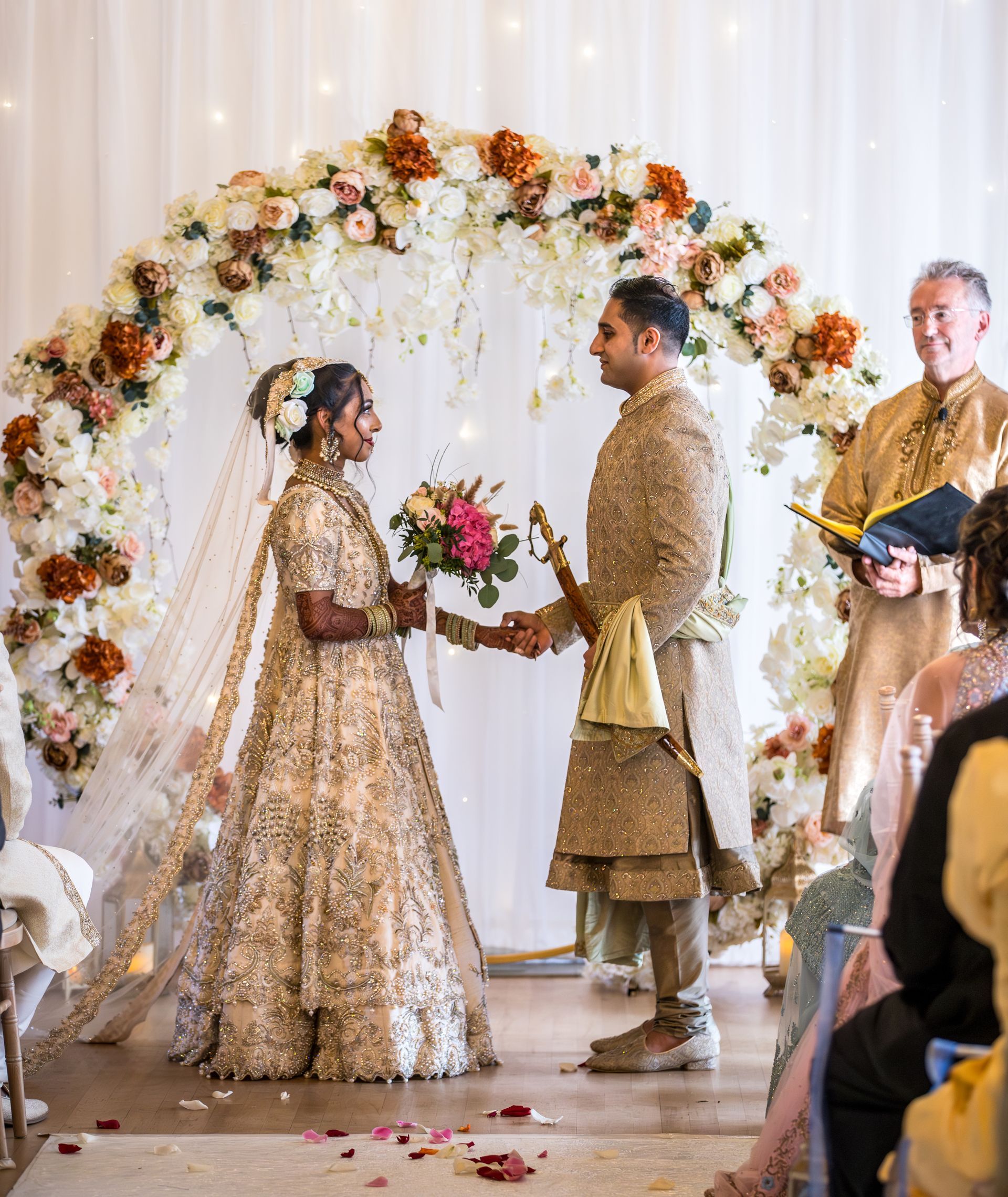 Asian bride and groom wearing gold and cream traditional dress holding hands looking into each others eyes at their wedding ceremony
