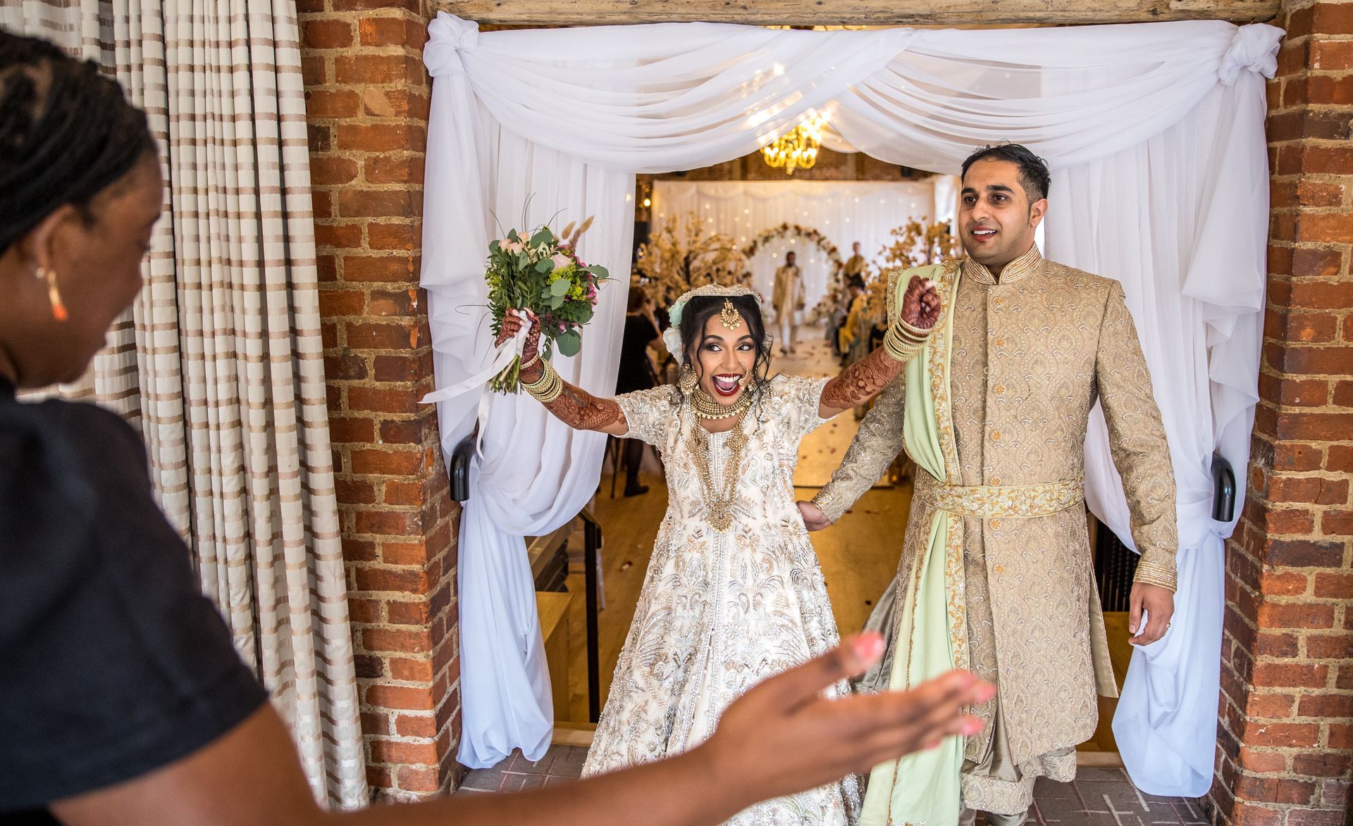 Indian bride in embroidered traditional cream and gold gown smiling with arms raised high holding bouquet