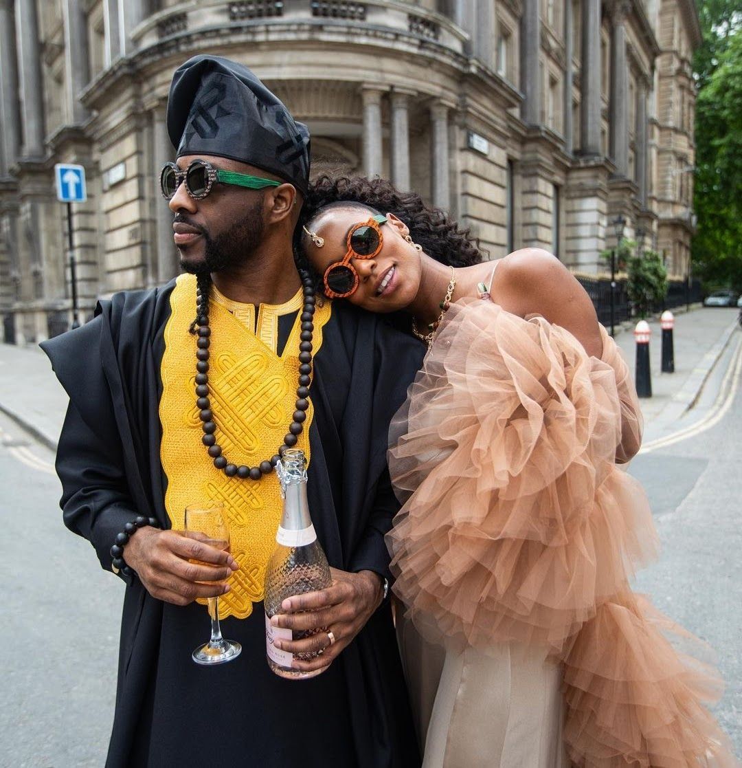 Black bride and groom for a multicultural wedding in traditional African outfits laughing and leaning on each other, standing in a London street