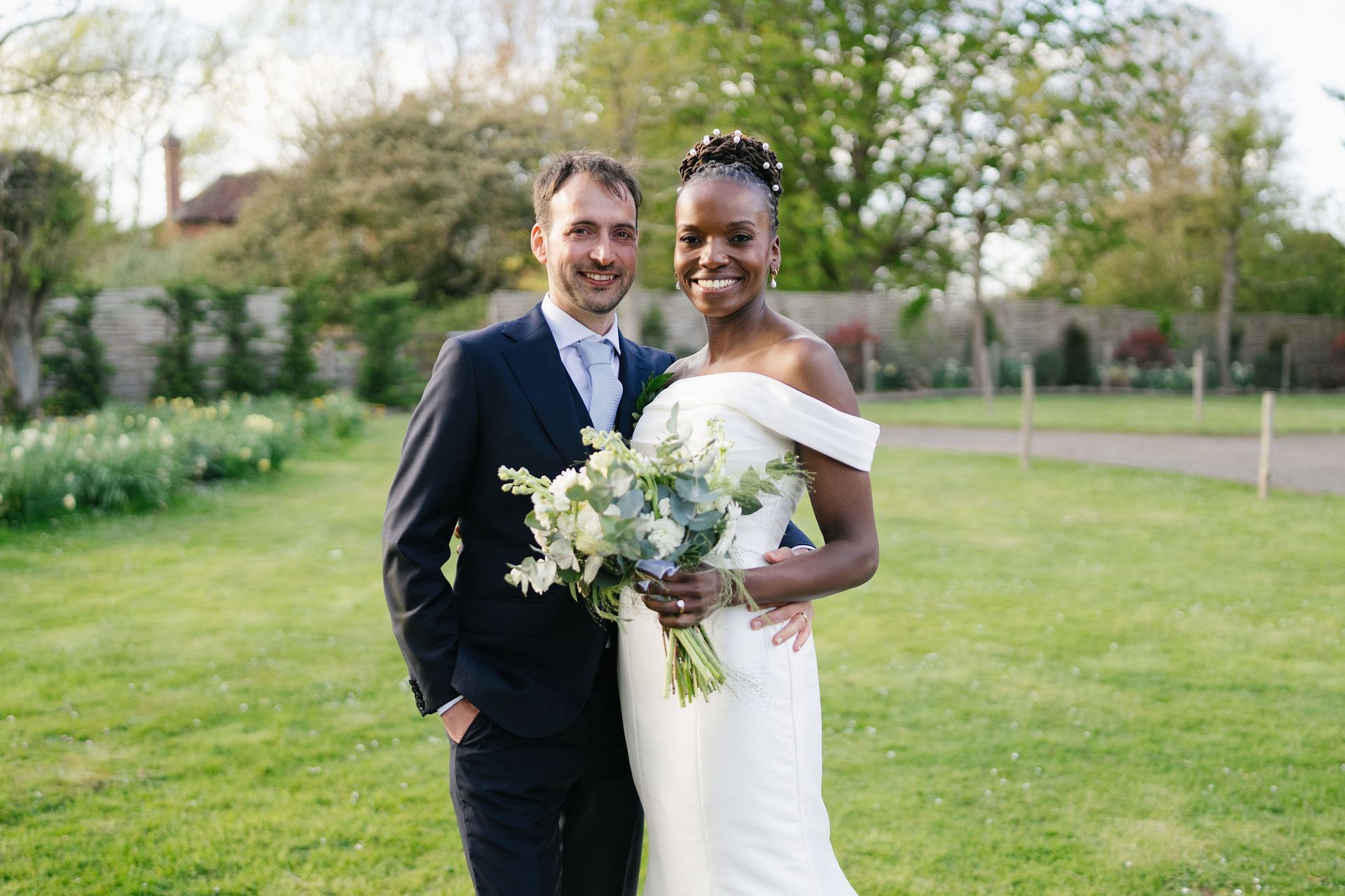 Black bride in white wedding dress holding bouquet with groom in blue suit beside her, in garden marquee wedding both smiling at the camera