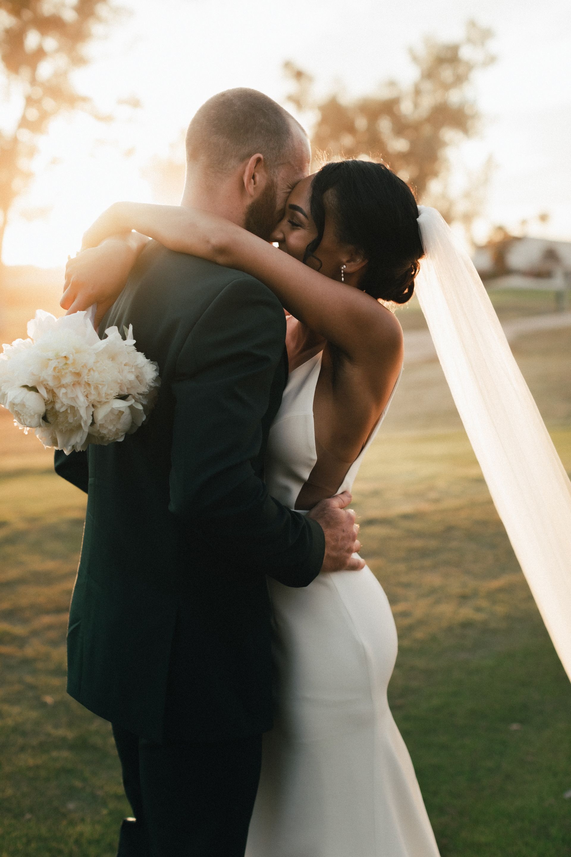 Black bride in white wedding dress holding a white flower bouquet and white male in dark green suit hugging each other outside